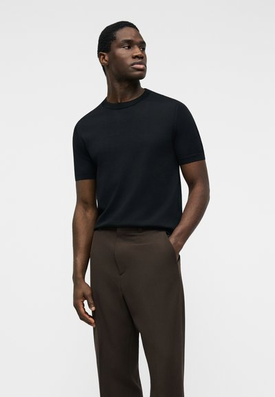 Άνδρας που φοράει απλό μαύρο κοντομάνικο πουκάμισο και καφέ παντελόνια, στέκεται με το ένα χέρι στην τσέπη, κοιτώντας προς τα πλάγια μπροστά σε απλό φόντο.