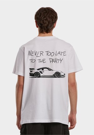 Person med kort brunt hår som har på seg en hvit T-skjorte med en svart sportsbil og teksten "ALDRI FOR SENT TIL FESTEN" på baksiden.