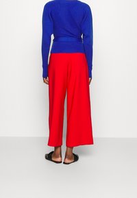 Osoba stojąca tyłem, ubrana w niebieską bluzę z długim rękawem, czerwoną szeroką spódnicę do kostek oraz czarne klapki bez zapięcia na jednolitym tle.