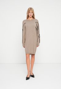 Robe tricotée beige à manches longues, ornée de broderies abstraites noires le long des manches. Texture douce, design mi-long, coupe décontractée.