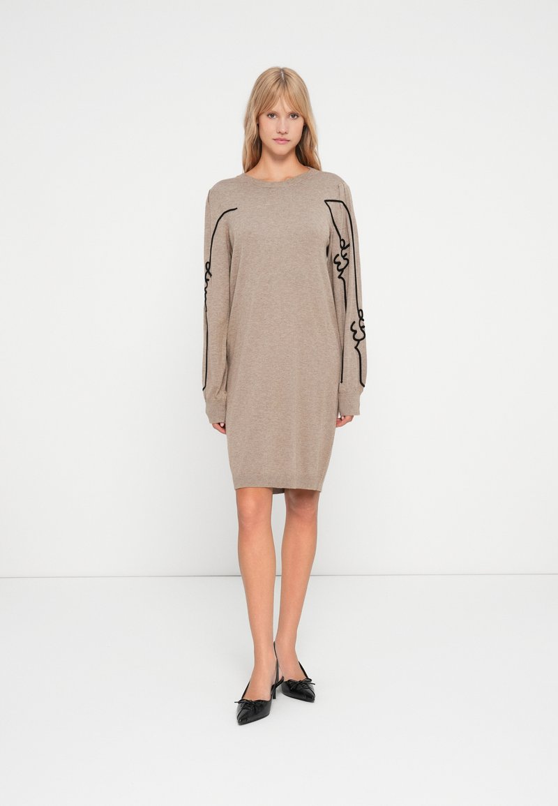 Robe tricotée beige à manches longues, ornée de broderies abstraites noires le long des manches. Texture douce, design mi-long, coupe décontractée.