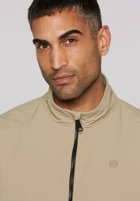 Light beige zip-up jacket with a high collar and a subtle logo on the chest. Features a smooth texture and black zipper detail.
