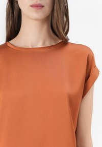 Koszulka z krótkim rękawem w kolorze rdzy pomarańczowej, wykonana z gładkiego, lekkiego materiału. Posiada okrągły dekolt oraz luźny krój z wykończonymi rękawami.