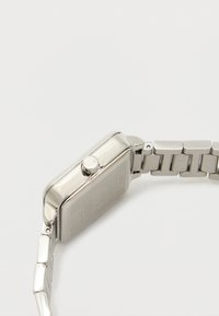 Hõbedane ristkülikuline kell tekstuurse kellasehitusega, õhuke äär ja tugev metallist linkriba, mille küljes on reguleerimine jaoks kroon.