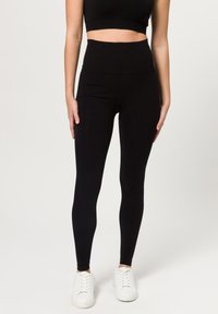 Leggings neri a vita alta con un tessuto liscio ed elasticizzato e una vestibilità aderente, abbinati a scarpe da ginnastica bianche.