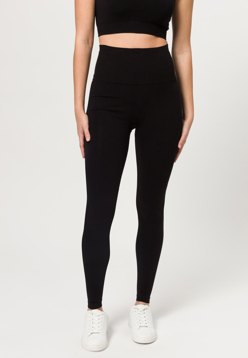Leggings neri a vita alta con un tessuto liscio ed elasticizzato e una vestibilità aderente, abbinati a scarpe da ginnastica bianche.