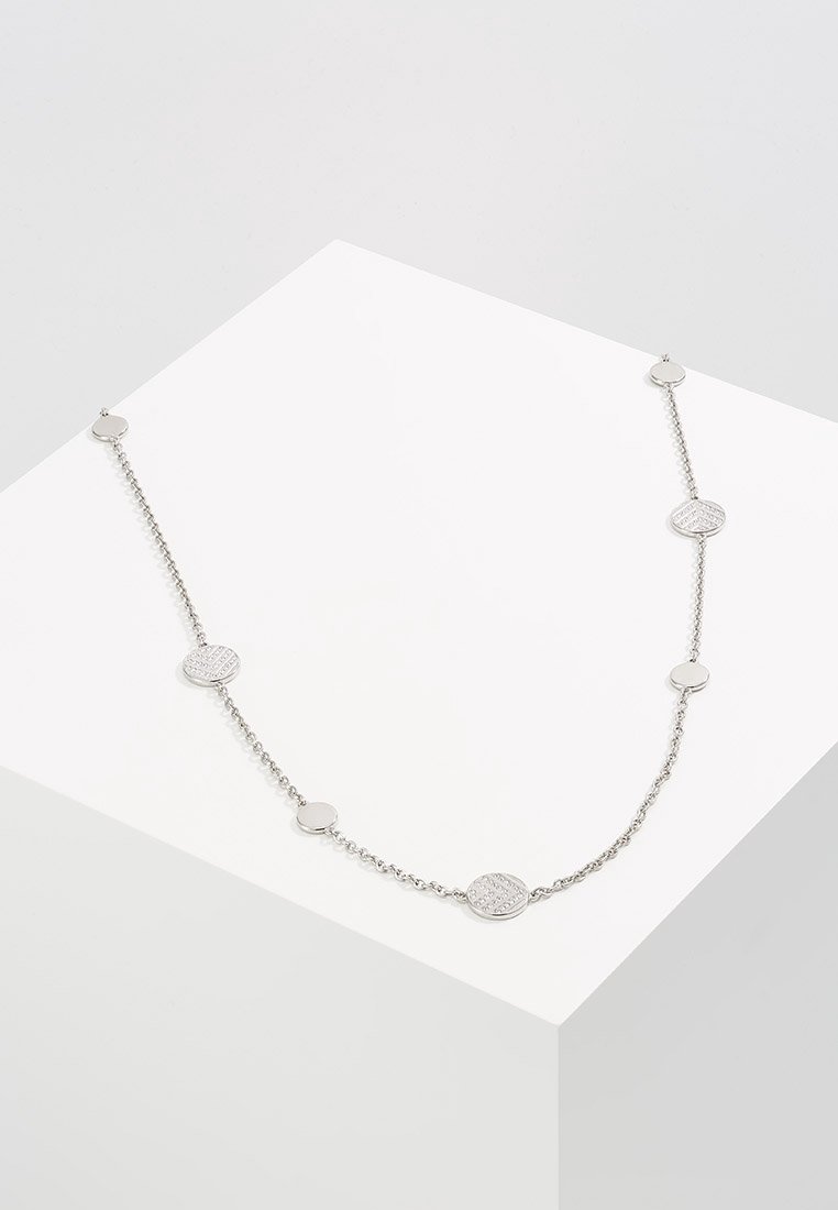 Collier chaîne en argent avec des disques ronds lisses et texturés alternés exposés sur un piédestal géométrique blanc.