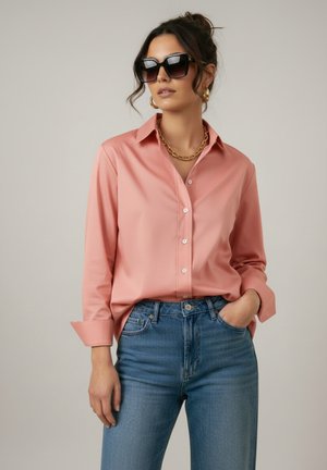 Vrouw draagt oversized zwarte zonnebril, gouden oorringen en kettingketting, roze overhemd met knopen en blauwe spijkerbroek met één hand in de zak.