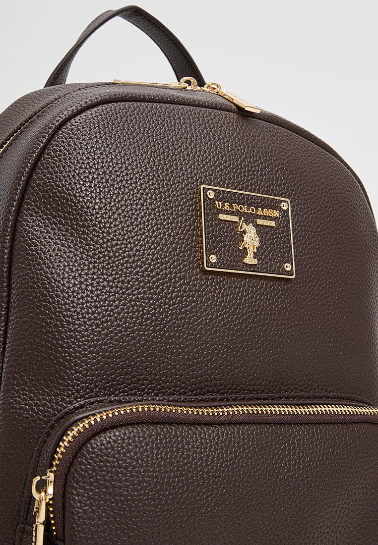 Brauner Leder-Rucksack mit strukturiertem Oberflächen, goldfarbenen Beschlägen und einem Logo-Schild. Enthält ein Reißverschlussfach für die Aufbewahrung.