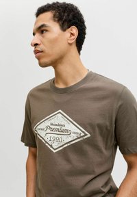 Hombre con cabello corto y rizado, vistiendo una camiseta marrón con el logotipo blanco vintage "Jack & Jones Premium 1990", mirando hacia la izquierda contra un fondo liso.