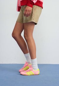 Rózsaszín sportcipők sárga és zöld részletekkel, sima dizájnnal, fűzővel és texturált talppal, bézs rövidnadrággal és fehér zoknival párosítva.