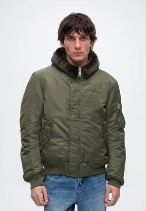 Homme portant une veste bomber vert olive avec un col en fourrure et un jean bleu, debout devant un fond blanc uni.