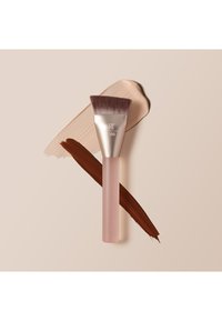 Pinceau de maquillage à poils biseautés reposant sur des échantillons de fond de teint beige et marron sur un fond neutre.