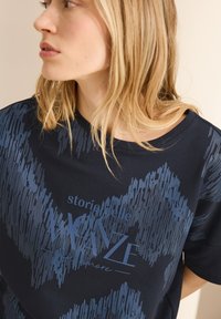 Camiseta de manga corta azul marino con un patrón abstracto texturizado en un azul más oscuro; presenta un texto impreso en azul claro en el centro.