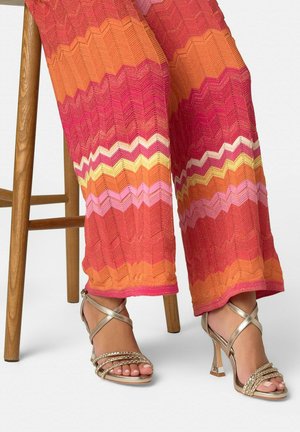 Personne assise sur un tabouret en bois portant un pantalon à motif zigzag multicolore et des sandales à talons métalliques avec des brides autour de la cheville.