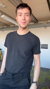 Nuori mies mustassa t-paidassa ja housuissa, seisomassa sisätiloissa, käsi taskussa ja lievä hymy kasvoillaan, teollisen katon taustaa vasten.