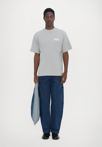 Camiseta gris de algodón con mangas cortas y un pequeño logo en el pecho, combinada con pantalones cargo de denim azul de corte holgado y zapatos negros.