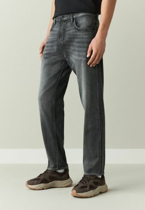 Man wearing faded black jeans, white socks, and dark brown chunky sneakers standing against plain background.