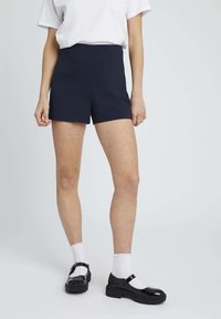 Des shorts taillés navy présentent une taille haute, un tissu lisse et une silhouette structurée. Associés à des chaussettes blanches et des chaussures en cuir vernies noires.