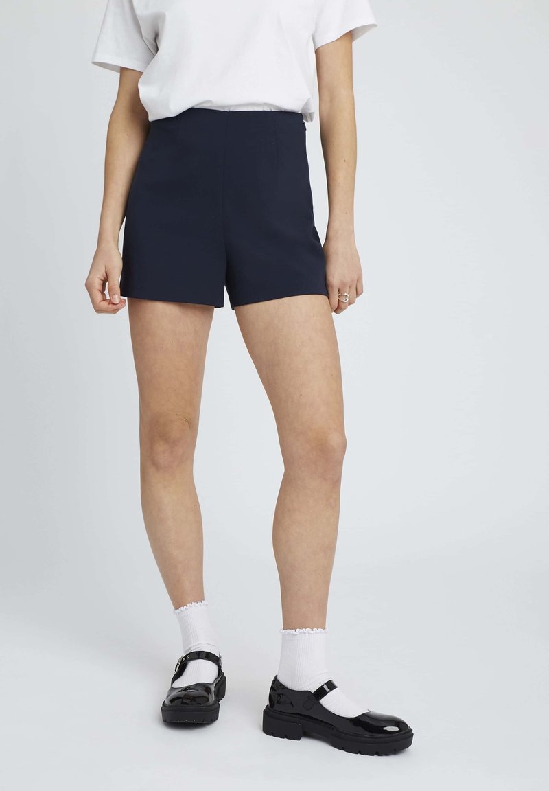 Des shorts taillés navy présentent une taille haute, un tissu lisse et une silhouette structurée. Associés à des chaussettes blanches et des chaussures en cuir vernies noires.