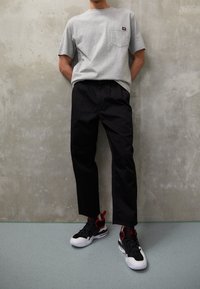 Homme portant une chemise gris à manches courtes, un pantalon noir et des baskets noires, blanches et rouges, debout devant un mur texturé gris.
