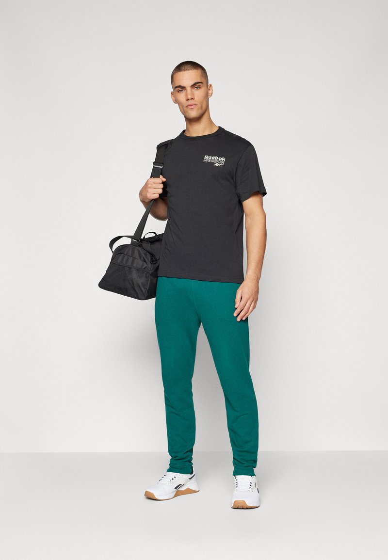 Camiseta negra con logo, pantalones de chándal verdes y zapatillas blancas. El modelo sostiene un bolso negro con correa para el hombro. Conjunto atlético casual en general.