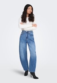 Blå jeans i vid passform med hög midja och lätt slitning, kombinerade med en figurnära vit tröja och svarta spetsiga högklackade skor.