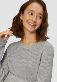 Grijze gebreide trui met een textuurpatroon, brede ronde halslijn en geribde mouwen, gecombineerd met een delicate gouden ketting.
