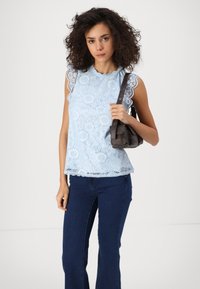 Ανοιχτό μπλε δαντελένιο τοπ με φαρδιά λαιμόκοψη και άκρες μανικιών σε σχήμα κυμάτου, συνδυασμένο με σκούρες μπλε φαρδιές τζιν παντελόνες; κρατώντας μια μικρή σκούρα τσάντα.