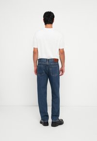 Jeans en denim bleu avec un design à jambe droite, des poches arrière et une étiquette en cuir marquée au niveau de la taille. Portés avec un t-shirt blanc uni.