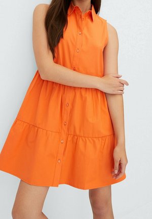 Femme portant une robe sans manches orange vif boutonnée avec une jupe à volants, debout, un bras croisé sur son corps.