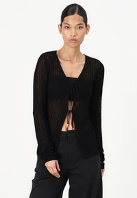 Cardigan nero lavorato a maglia con maniche lunghe, scollatura a V, chiusura con fiocco frontale e texture a coste. Indossato sopra pantaloni neri.