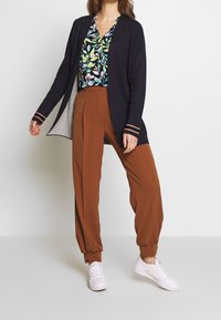 Marineblaue Strickjacke mit orange gestreiften Bündchen, kombiniert mit einer gemusterten grünen Bluse mit Blumenmuster und braunen, weit geschnittenen Hosen, abgerundet durch weiße Sneakers.