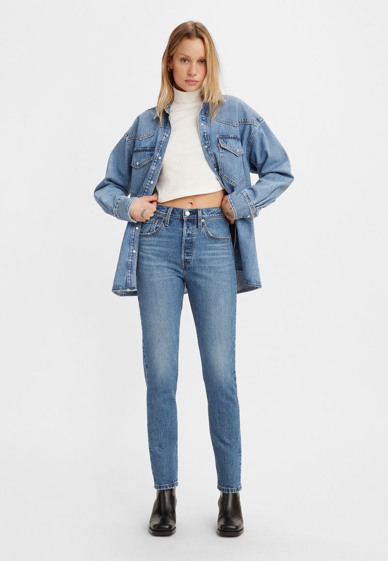 Helles blaues Denim-Jacket über einem weißen, cropped Rollkragenpullover, kombiniert mit hellblauen, hochgeschnittenen Jeans und schwarzen Stiefeletten.