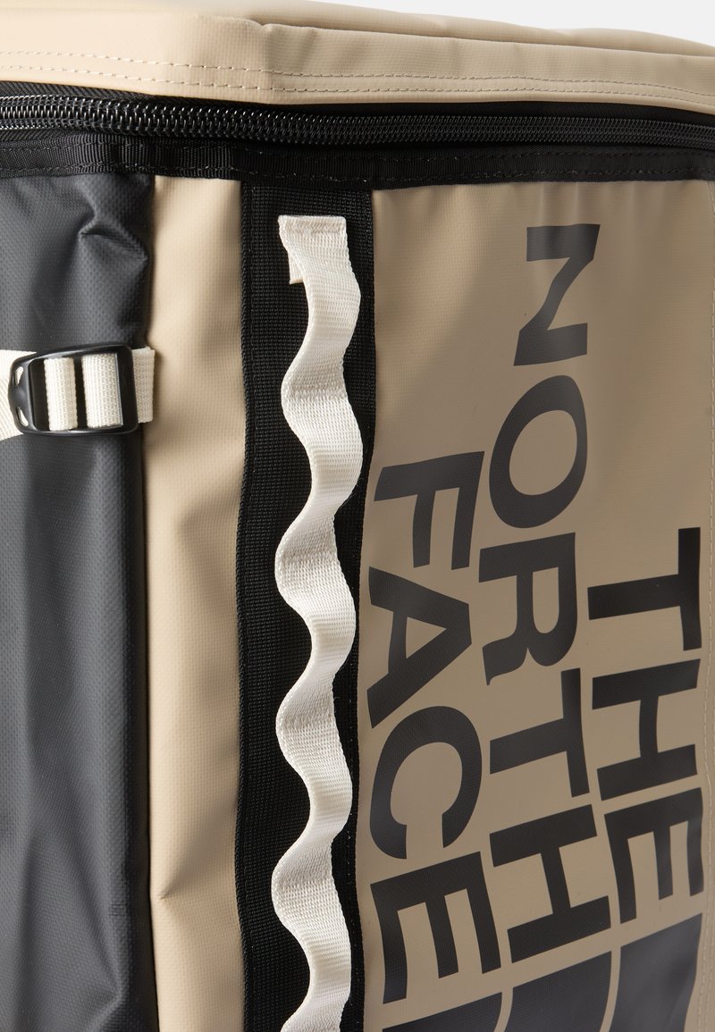 Isoleret køletaske i beige og sort med et stort logo, tekstureret stof og hvide hardware-detaljer, samt en zigzag-rem.
