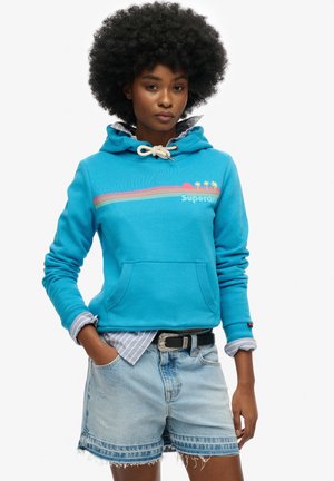 Jeune femme aux cheveux afro portant un sweat à capuche bleu imprimé de palmiers, superposé à une chemise rayée, et un short en jean.