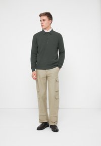 Polo en maille vert foncé avec trois boutons, associé à un pantalon cargo beige avec des poches latérales et des poignets retroussés. Des chaussures noires complètent le look.