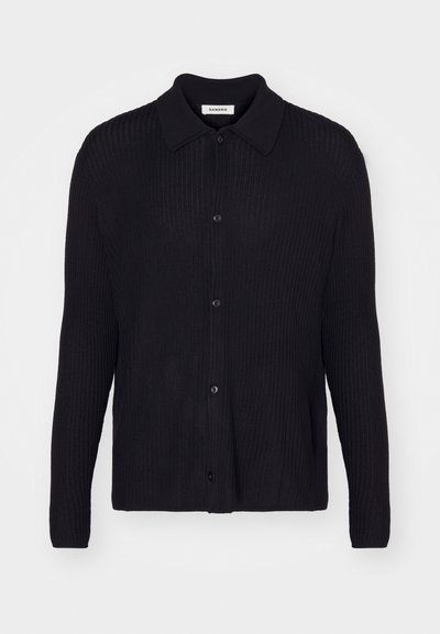 Svart ribbad stickad kofta med långa ärmar, knappstängning fram och spetsig krage på en enfärgad bakgrund.