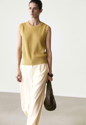 Donna che indossa un top senza maniche color senape a maglia e pantaloni larghi color crema, con una borsa verde scuro, in piedi davanti a uno sfondo bianco semplice.