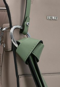 Dettaglio di una borsa a mano in pelle beige con una tracolla in pelle verde avvolta attorno a un anello di metallo argento. Texture liscia con cuciture a contrasto.