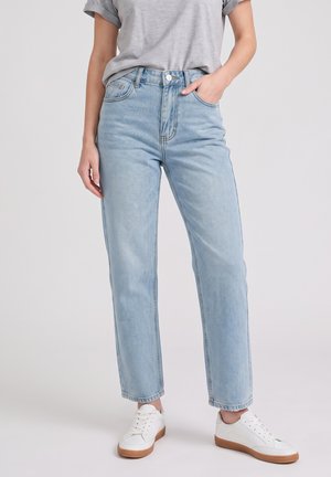 Personne portant un jean droit taille haute bleu clair, un t-shirt gris à manches courtes retroussées, des baskets blanches avec des semelles marron, une main dans la poche.