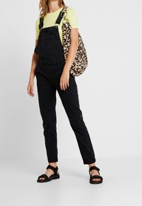 Svarta denimsoveraller med justerbara remmar, i kombination med en neongul ribbad topp och en väska med leopardmönster. Svarta sandaler fulländar stilen.