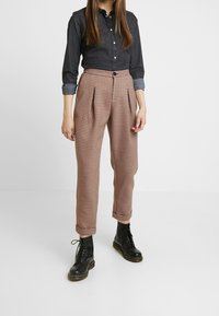 Černá džínsová košile s knoflíkovým zapínáním, spárovaná s hnědými kostkovanými kalhotami s sklady a ohrnutými manžetami, obutá do černých šněrovacích bot.