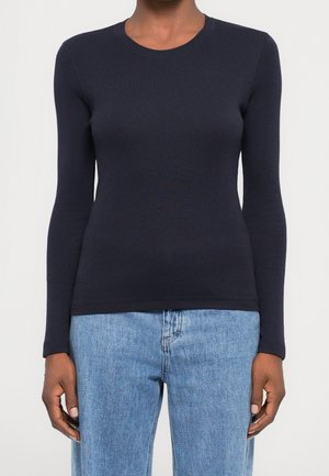 Osoba ubrana w dopasowany, granatowy top z długim rękawem oraz jasnoniebieskie, wysokie spodnie dżinsowe, stojąca na tle jednolitego, jasnego tła.