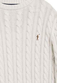 Pull en maille torsadée de couleur crème avec un col côtelé et une petite figure brodée marron jouant d'un cor sur la poitrine.