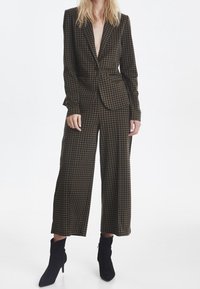 Kvinna i dräkt av brunt och svart hounds-tooth mönster med vida, korta byxor och matchande kavaj, i kombination med svarta spetsiga ankelskor.