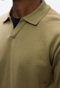 Homme portant un polo à manches longues vert olive par-dessus un haut noir à col rond, montrant le col et la partie supérieure de la poitrine.