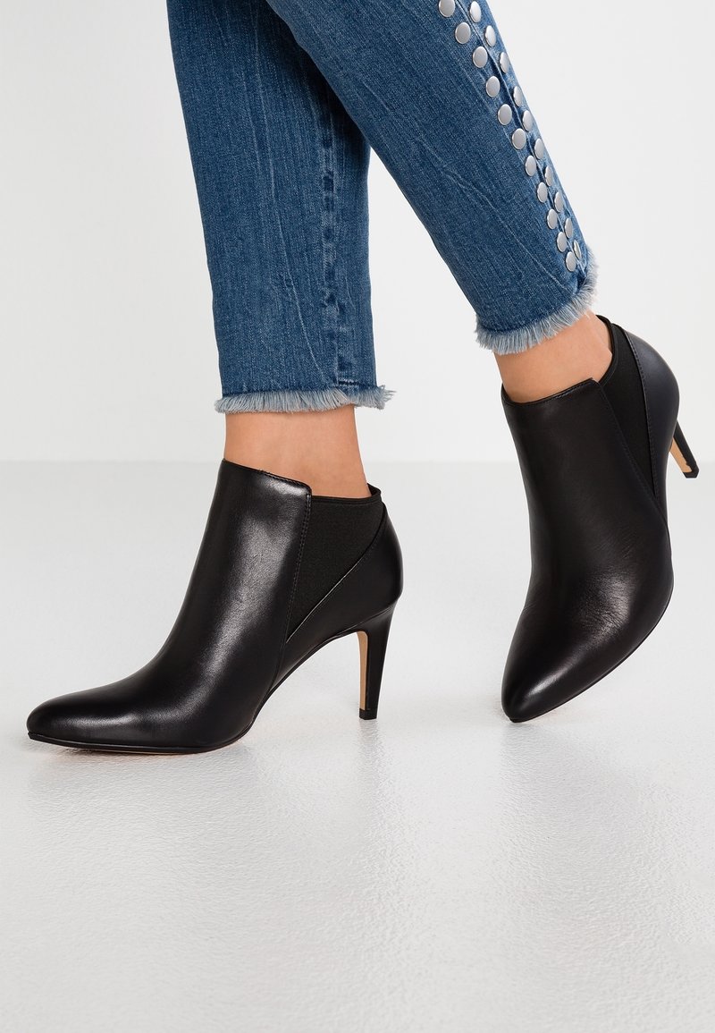 Botas de cuero negras hasta el tobillo con puntera afilada, tacón de aguja y paneles laterales elásticos. Llevadas con jeans de mezclilla con dobladillo deshilachado y detalles plateados.