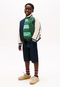 Χαμογελαστός αγόρι που φοράει πράσινο και μπλε varsity μπουφάν, πράσινο ριγέ πουλόβερ, σκούρα τζιν σορτς, ριγέ κάλτσες και μπεζ αθλητικά παπούτσια, στέκεται μπροστά σε λευκό φόντο.