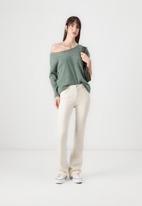 Grønn strikket av-shoulder genser, kremfargede utsvingte jeans, hvite joggesko, modell som holder en olivengrønn veske. Enkel, avslappet antrekk.